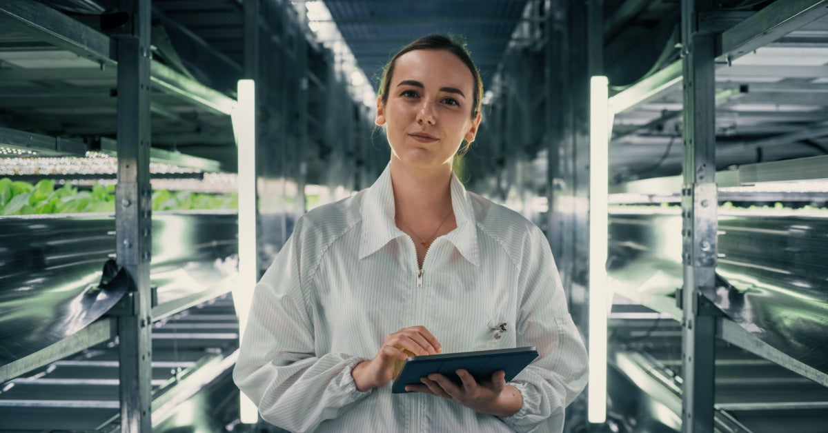 An agricultural engineer smiles as she stands in an aisle between two shelves of vertical grow racks with LED lights.